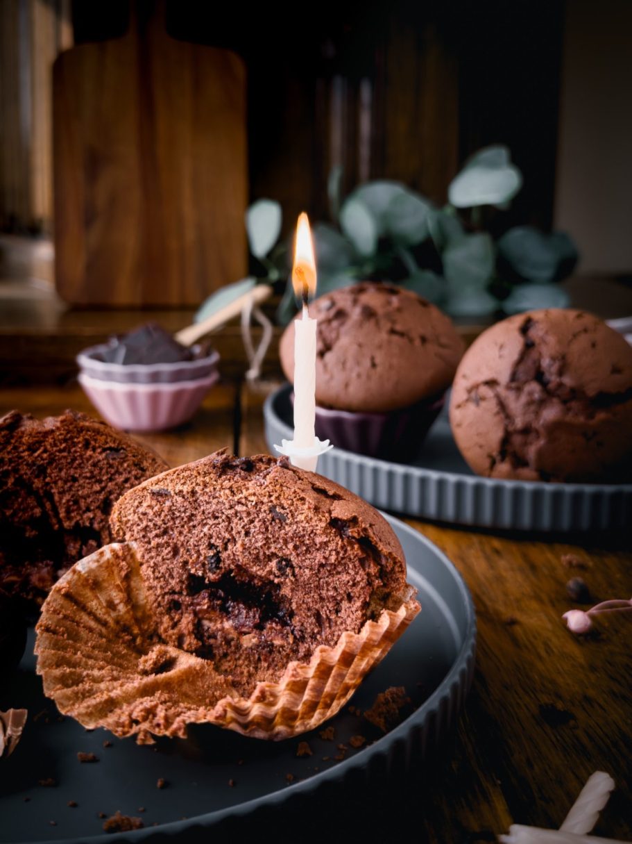 Foodfotografie Schokoladenmuffin Zwei Schokoladenmuffins sehen auf einem Teller. Dritter aufgeschnitten Muffin mit brennenden Kerze steht im Vordergrund auf zweitem Teller.