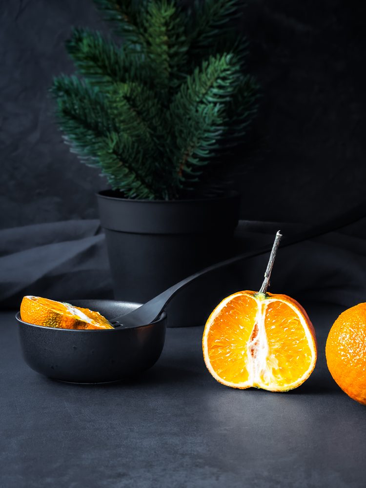 Dark & Moody Foto Durch die Mitte durchgeschnittene halbe Mandarine steht vor Tannengrün im Dank & Moody Style.