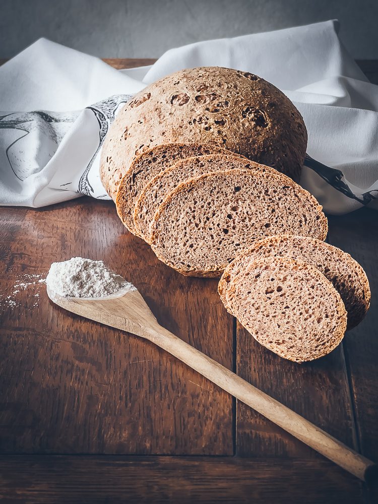 Foodfotografie Brot Auf einem Holztisch liegen aufgeschnittene Brotscheiben, außerdem eine weiße Serviette und ein Holzlöffel gefüllt mit Mehl.